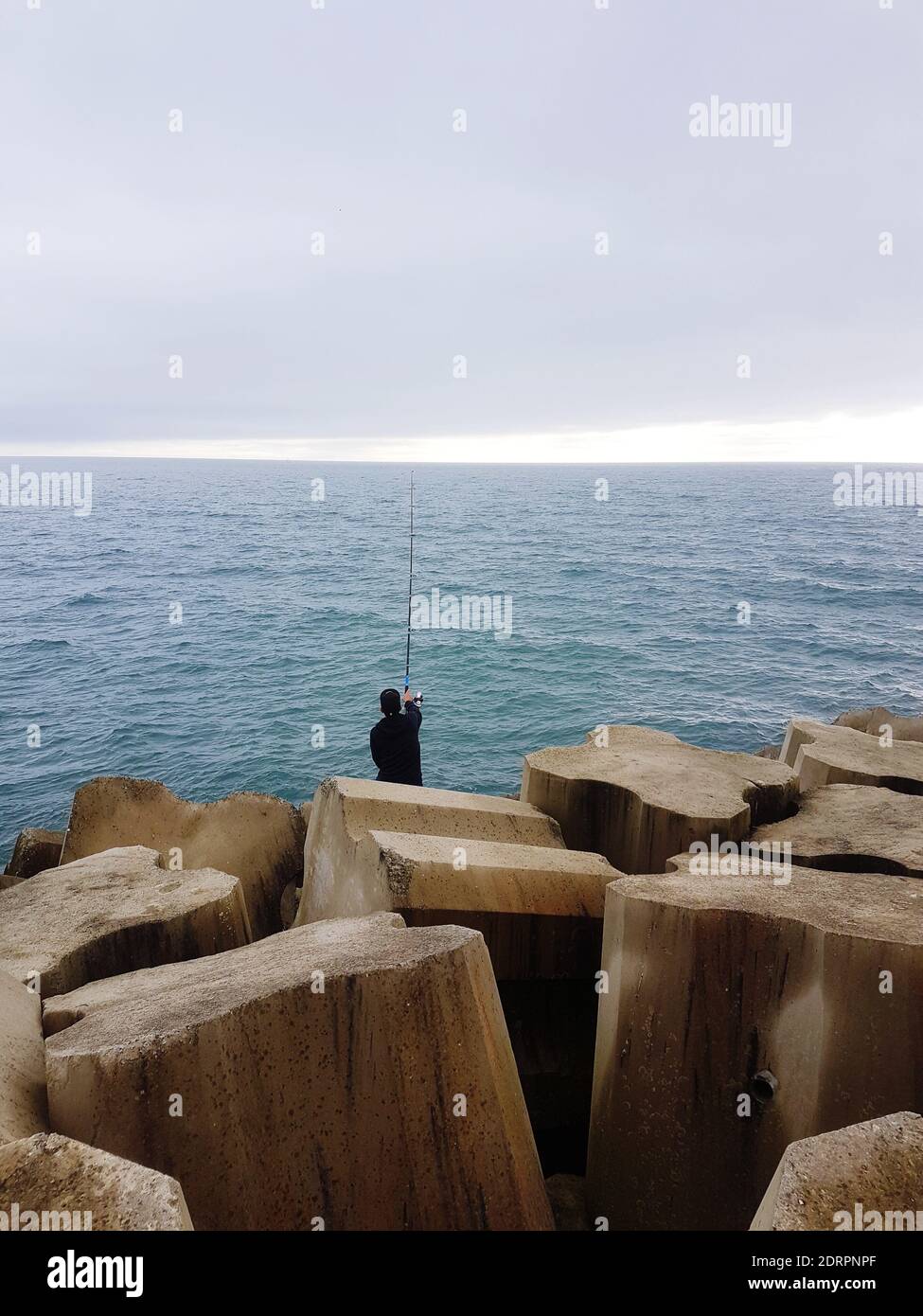 Rear View Of Mature Man Fishing In Sea While Sitting On Rock Against