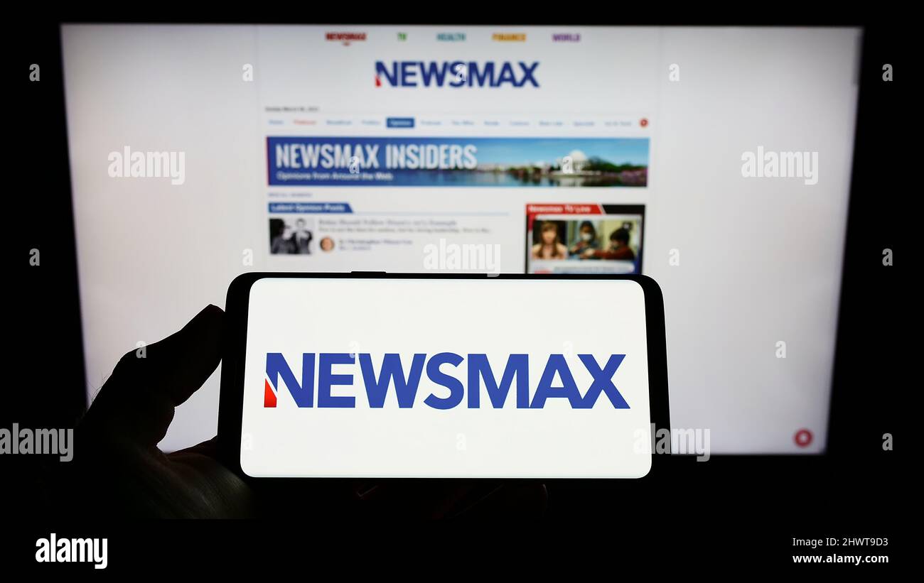 Person Holding Cellphone With Logo Of US Media Company Newsmax Media