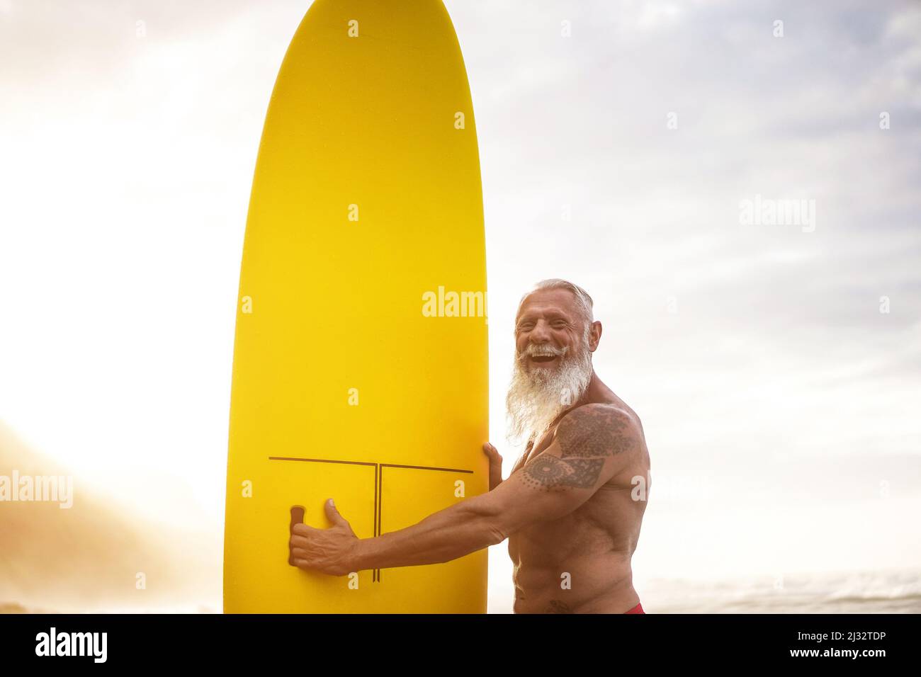 Happy Senior Surfer Man Holding Surf Board On The Beach At Sunset