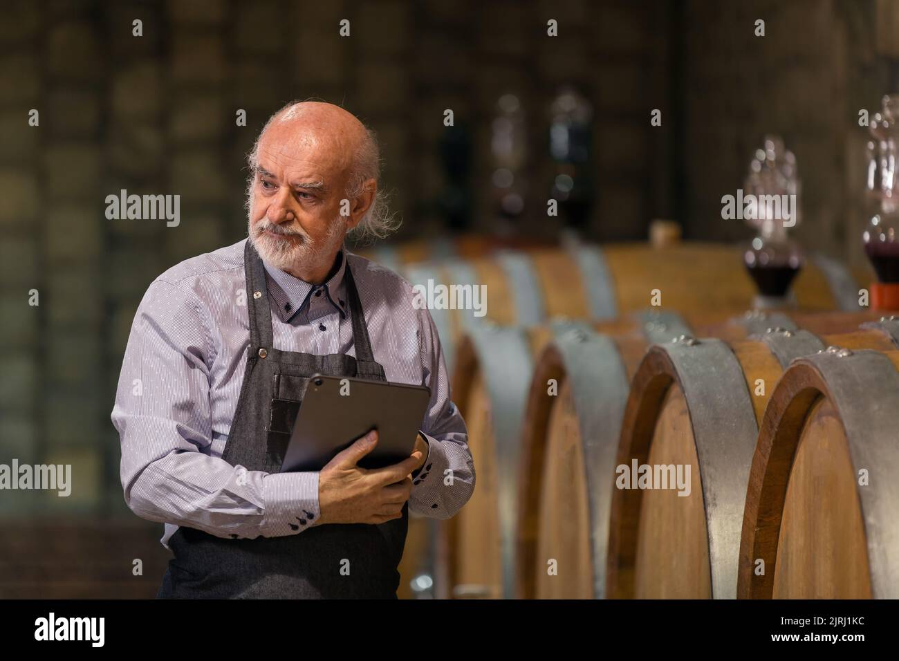 Senior Man Winemaker At Winery Checking Barrels In Wine Cellar Stock