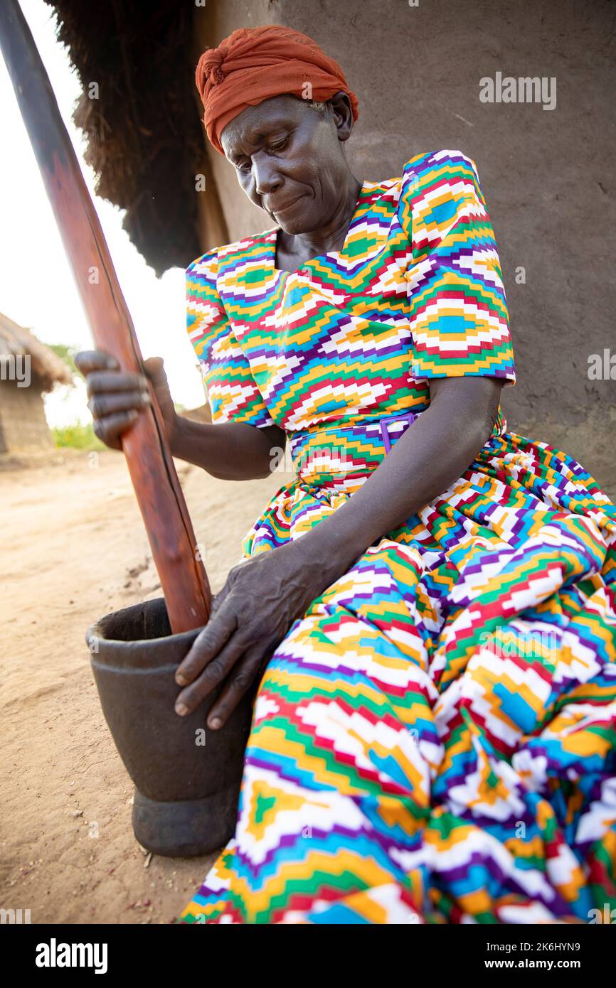 Elderly Woman In Traditional Colorful Dress Pounding Peanuts With A