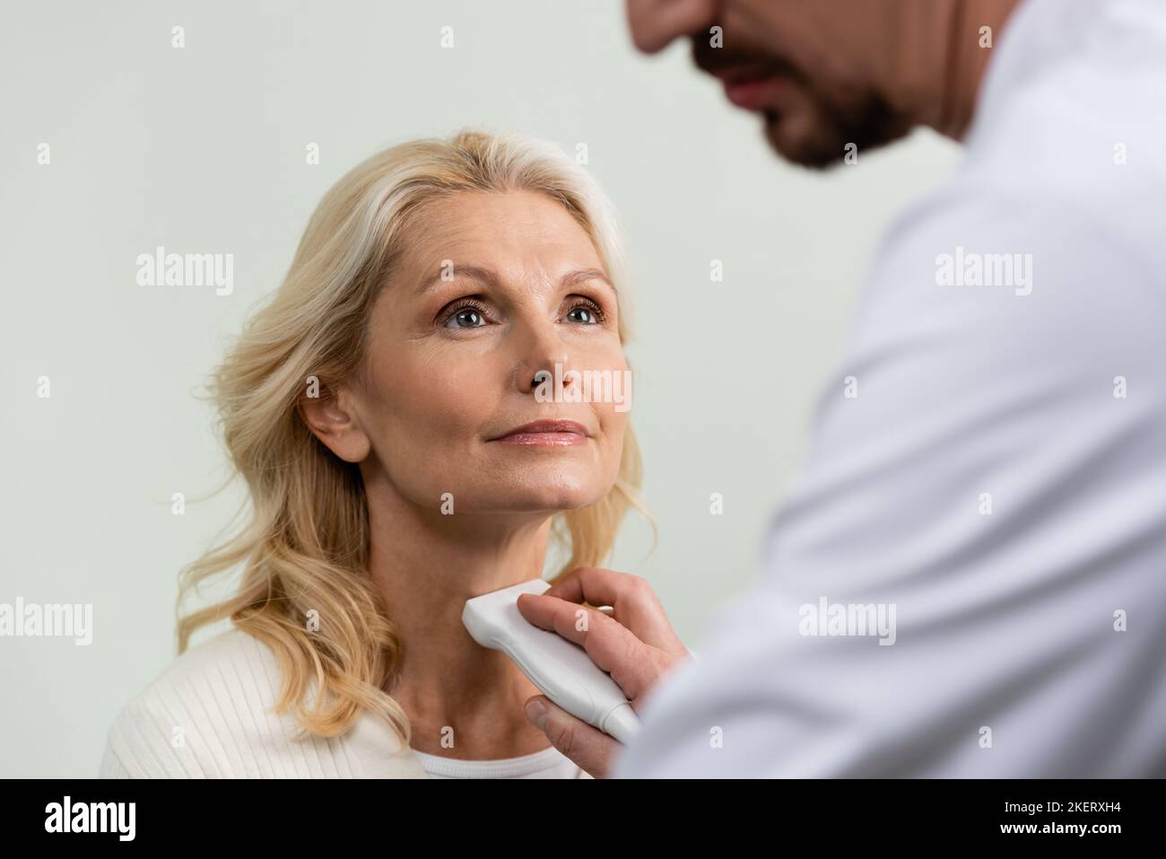 Blonde Woman Looking At Blurred Doctor Examining Her Throat With Ultrasound Stock Image Stock