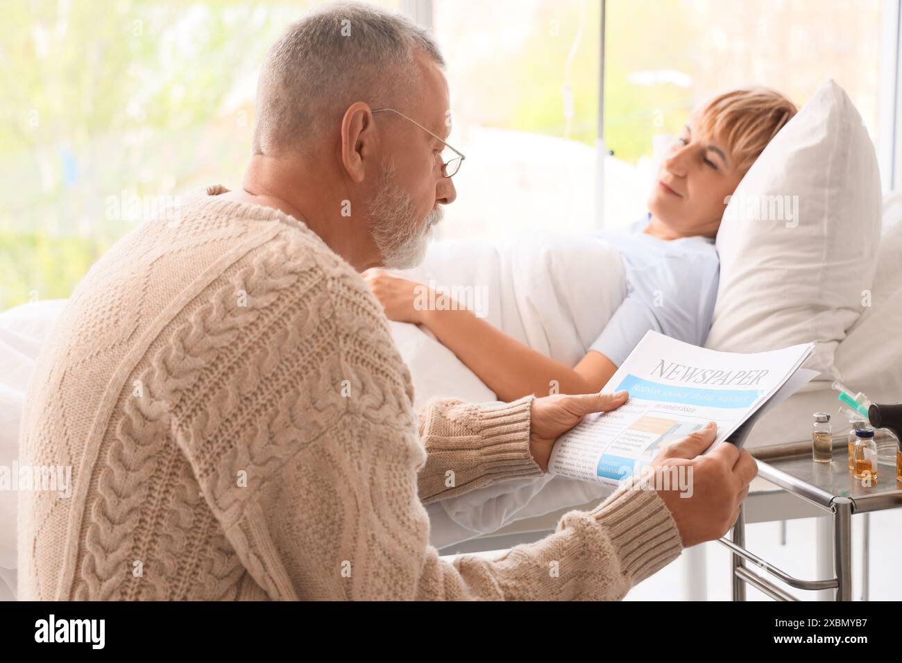 Mature Man Reading Newspaper With His Sick Wife Lying In Bed At