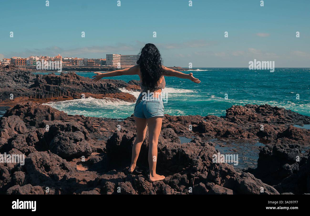 Two Women Stand On Lava Rocks Wearing Shorts And Bikini Tops With Their Arms Outstretched And