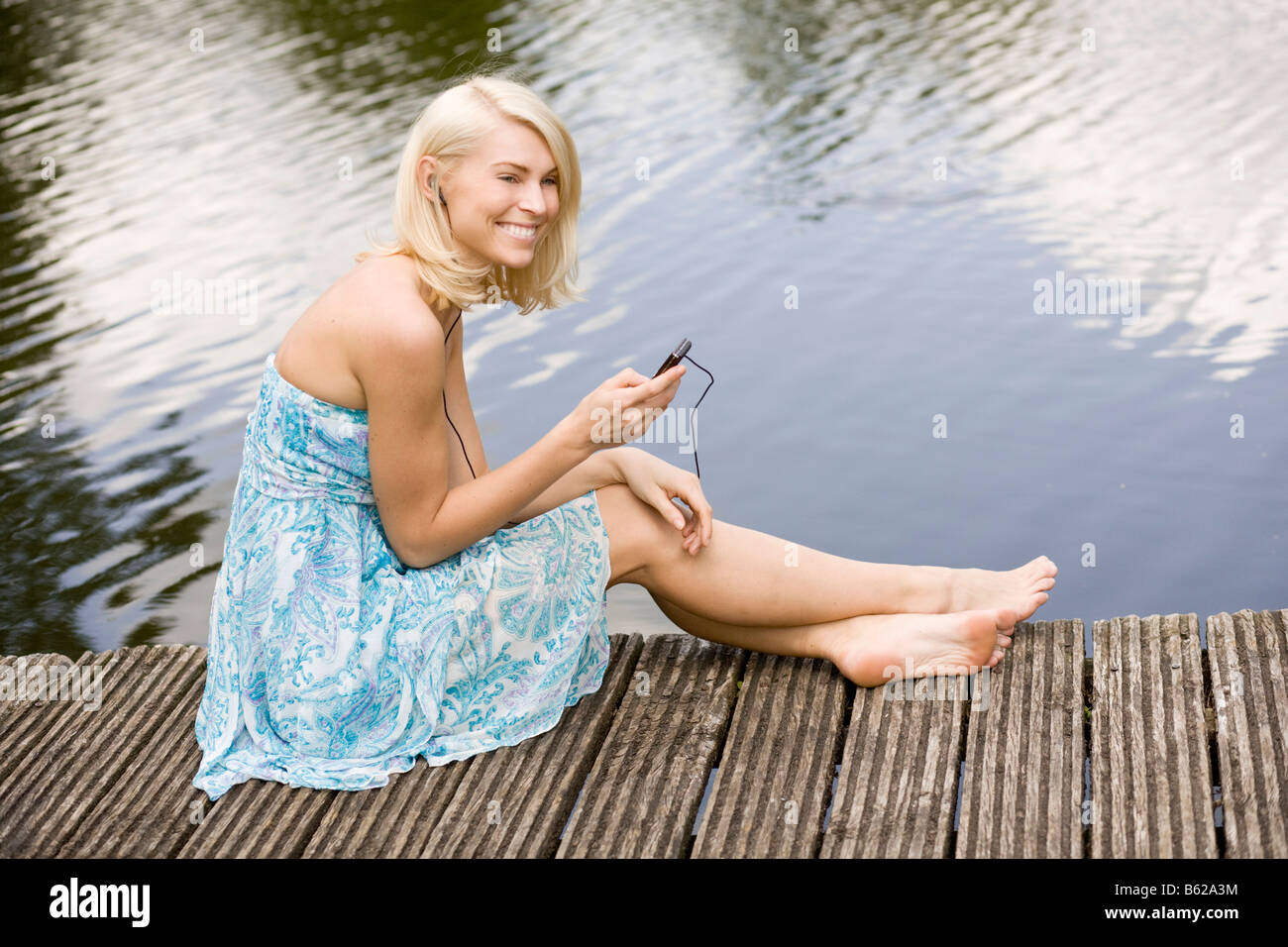 Young Blonde Woman Wearing A Dress While Sitting On A Wharf At A Lake