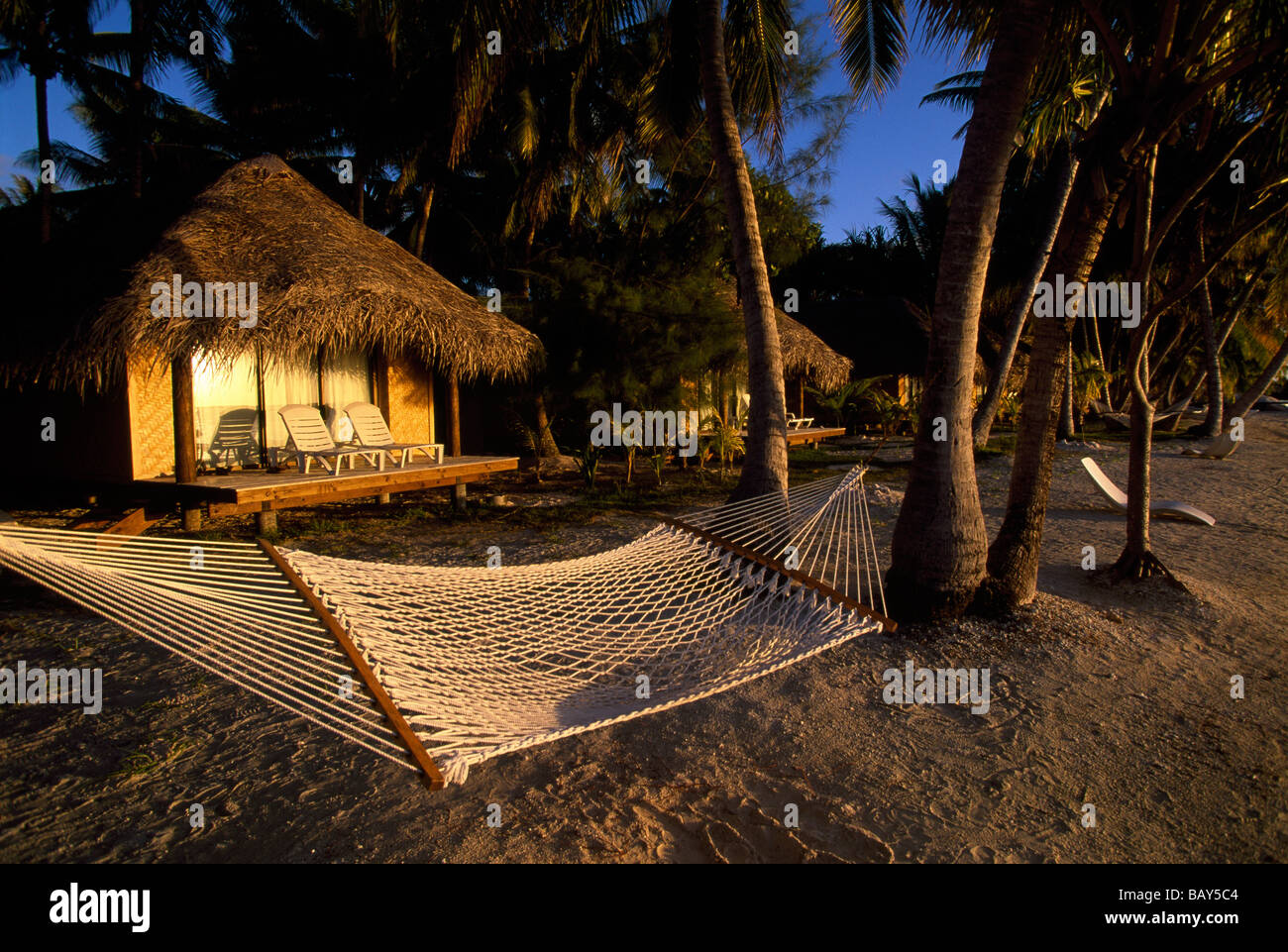 Hammock Between Palm Trees In Front Of Beach Huts In The Evening Sun