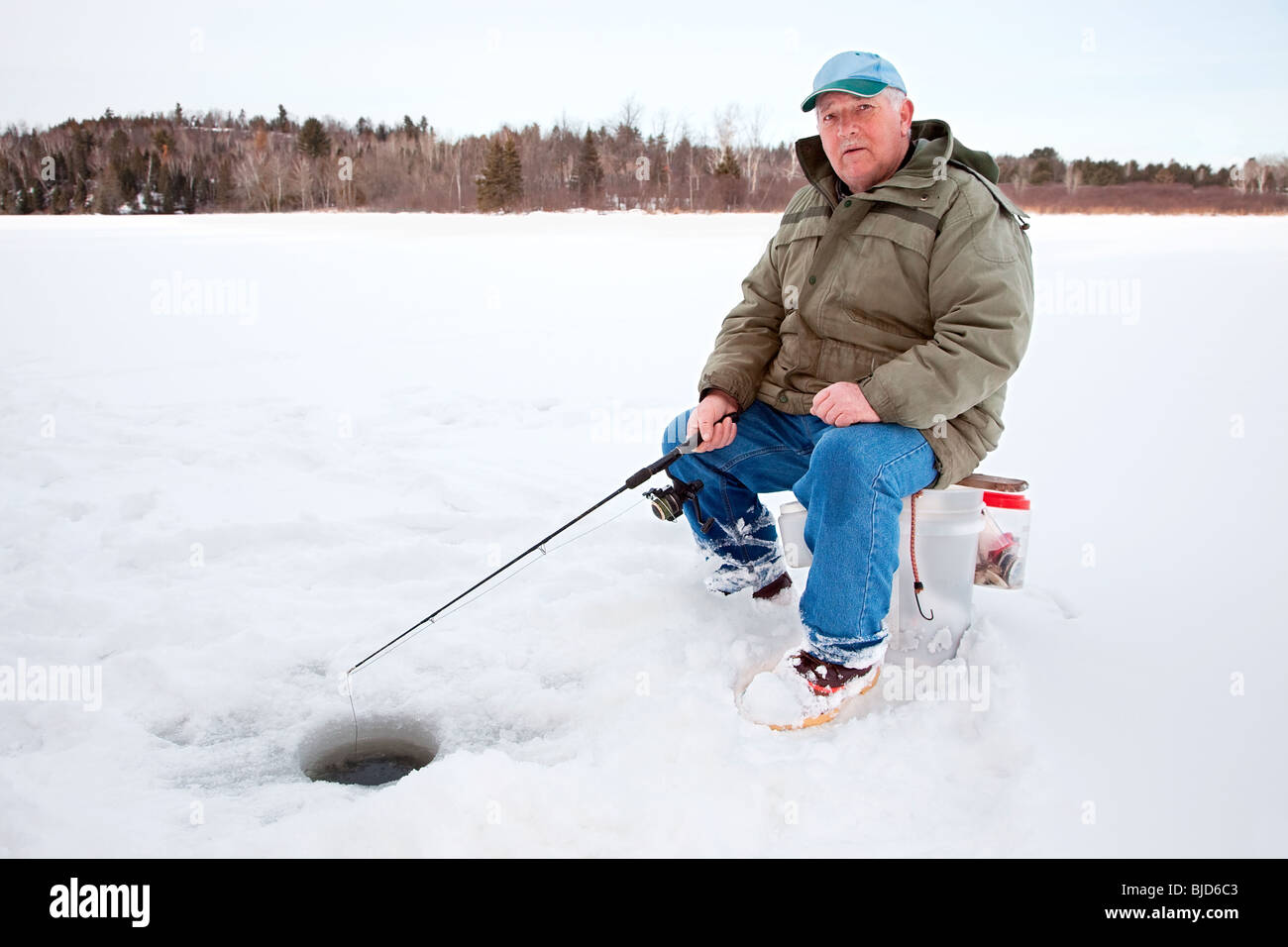 A senior man is ice fishing on the lake while sitting on a bucket Stock