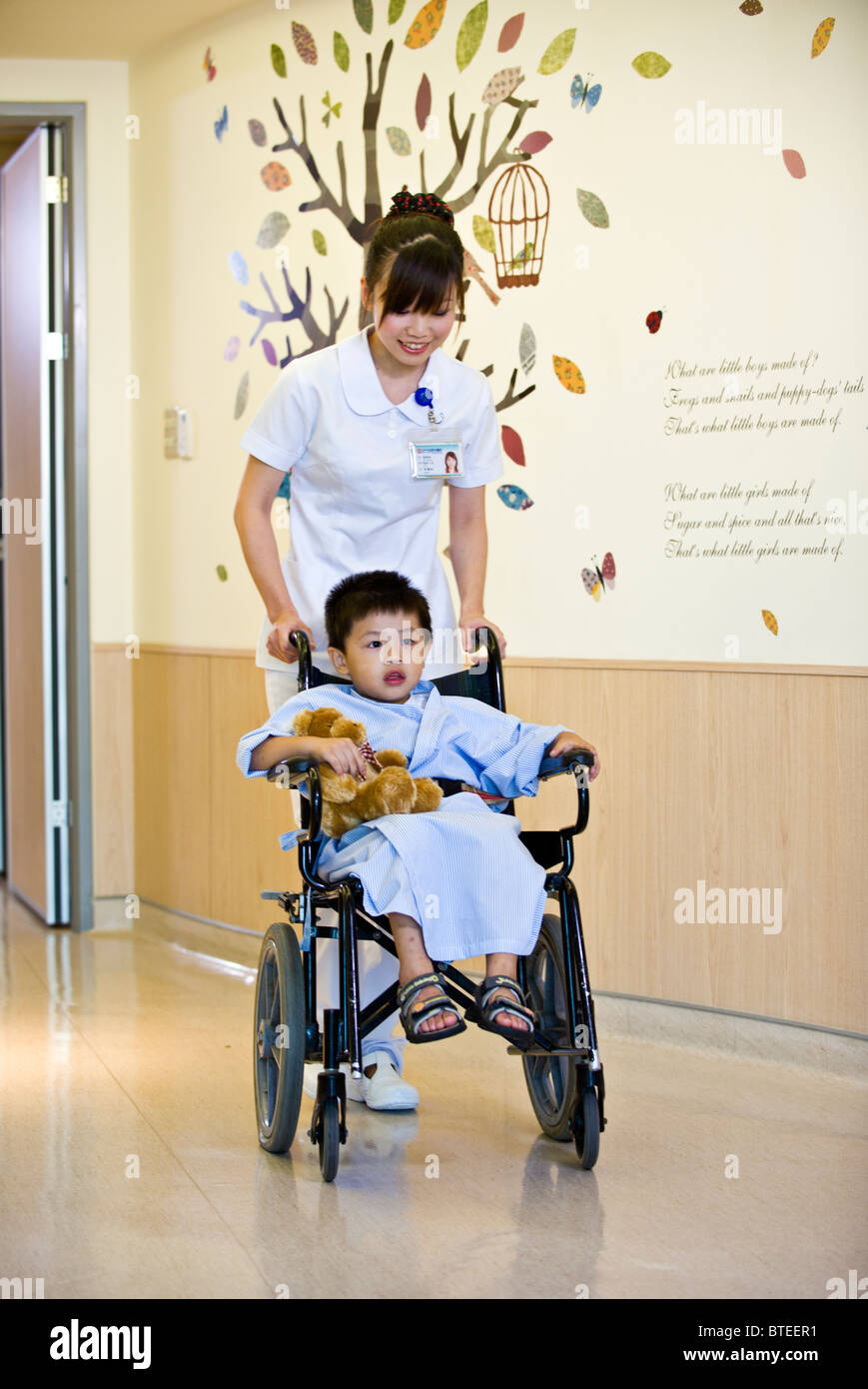 Nurse pushing young child patient in wheelchair down corridor in Stock