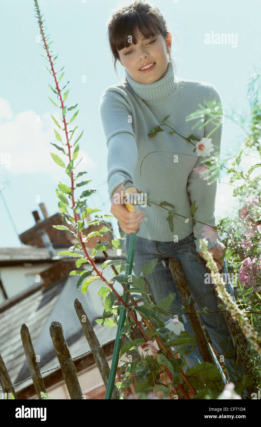 Emale With Brunette Hair Wearing Blue Poloneck And Jeans Leaning Over Fence Watering Garden With