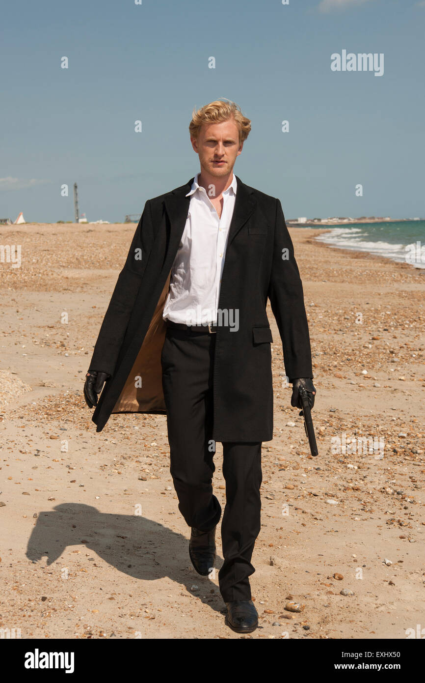 man in a long black coat holding a gun and walking on the beach ...