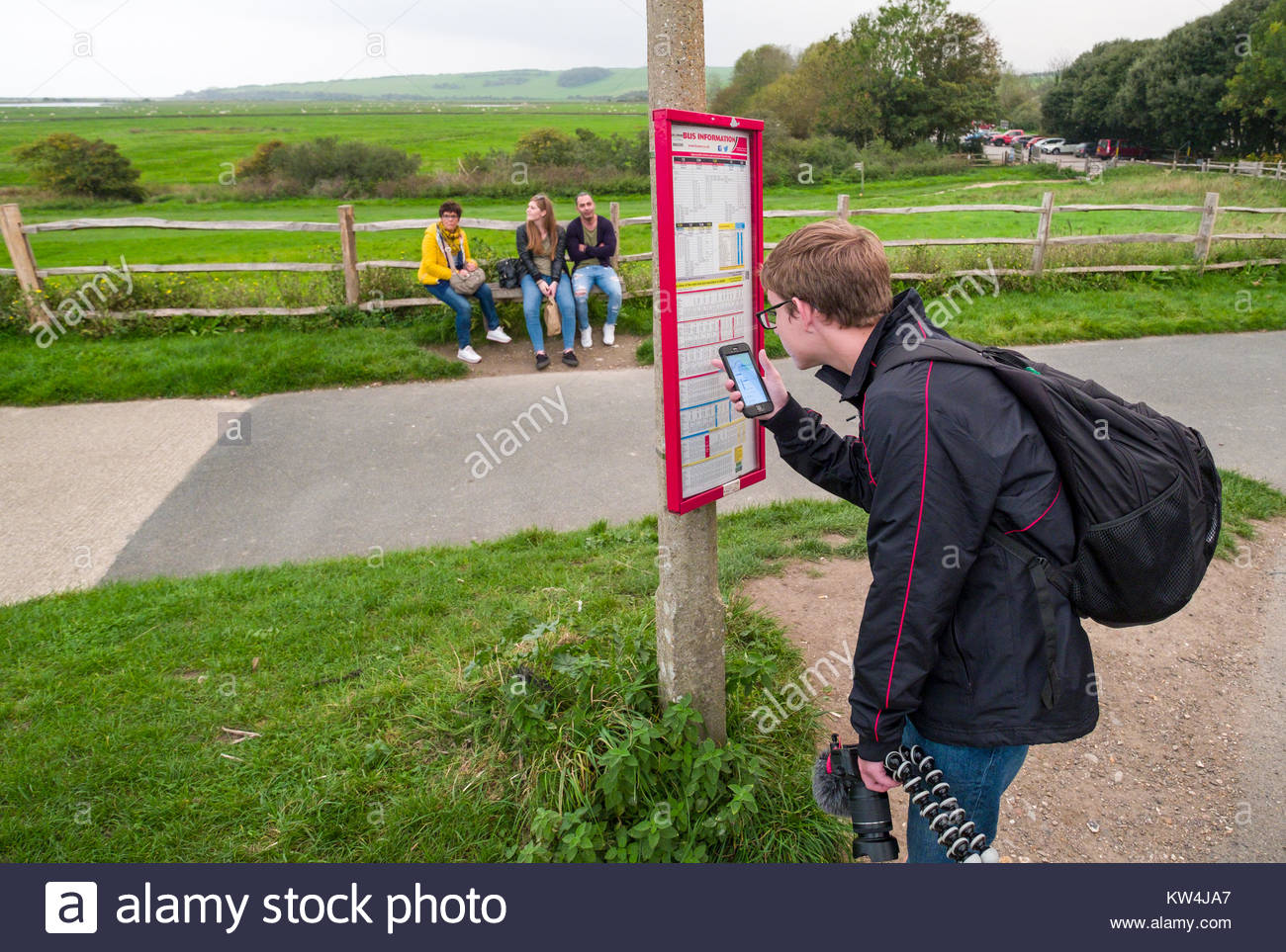 Young Man Checking Bus Schedule On His Smartphone Comparing To Posted Schedule At Bus Stop Exceat Cuckmere Haven Lewes District East Sus Engla Young Man Checking Bus Schedule On His Smartphone Comparing To Posted Schedule At Bus Stop Exceat Cuckmere Haven Lewes District East Sus Engla