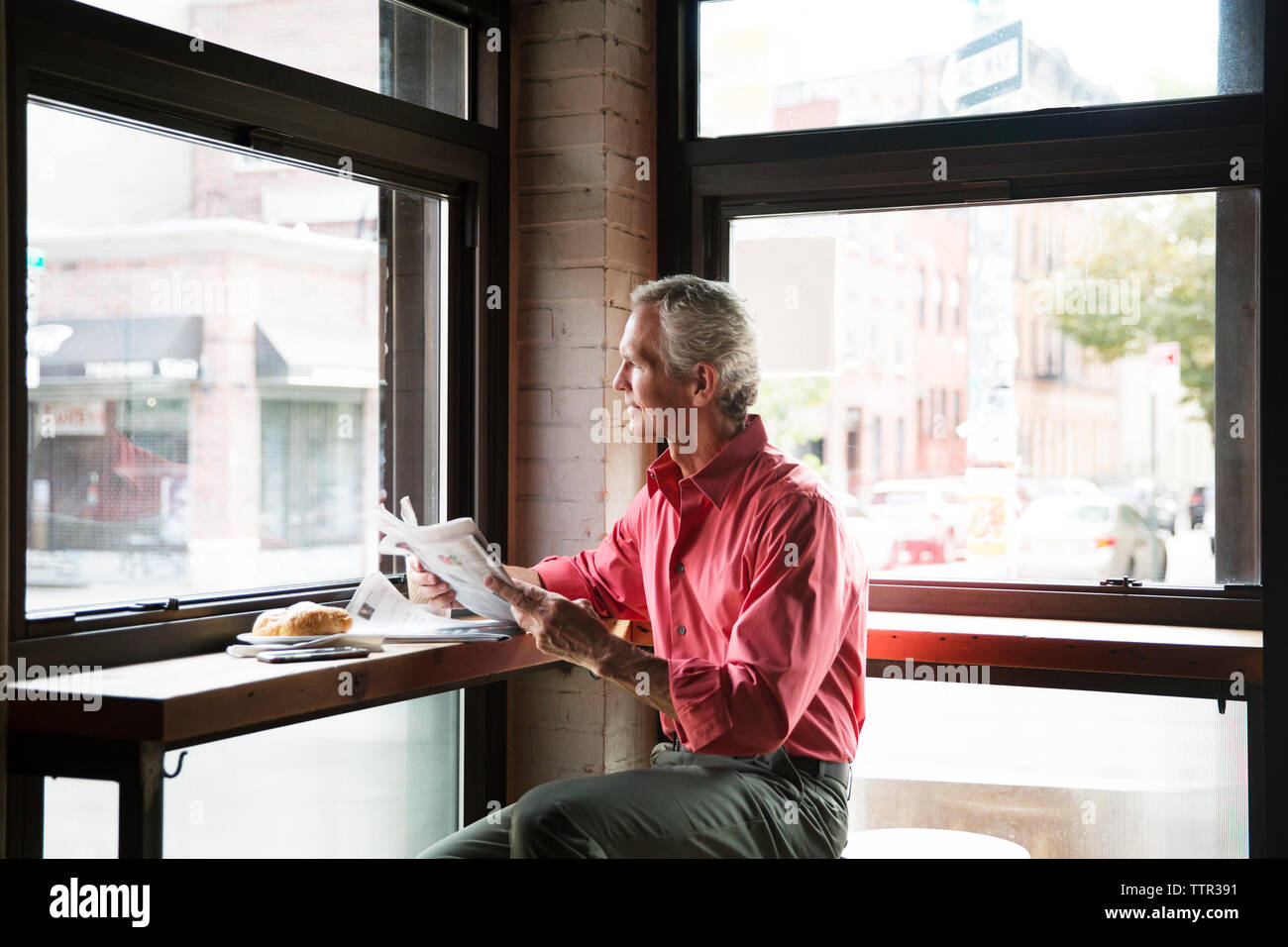 Mature Man Reading Newspaper While Having Croissant At Cafe Stock Photo