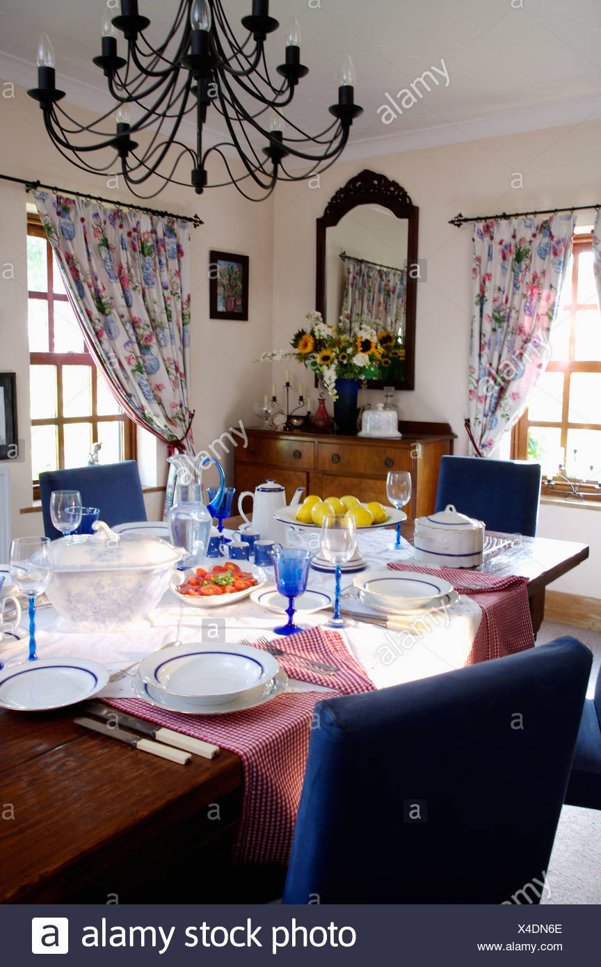 Wrought Iron Chandelier Above Blue Chairs And Table Set For Lunch With White Crockery Glasses In Country Dining Room Wrought Iron Chandelier Above Blue Chairs And Table Set For Lunch With White Crockery Glasses In Country Dining Room