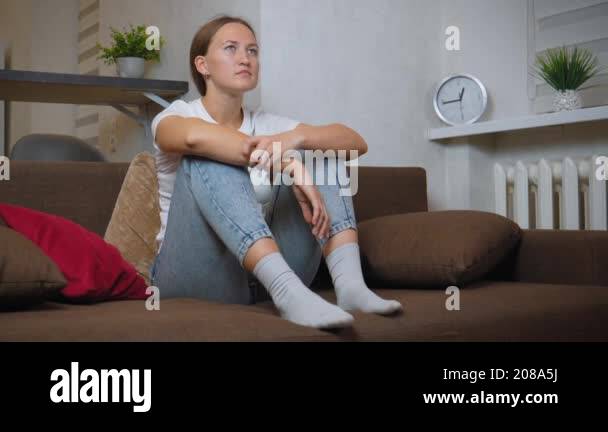 Young woman is sitting on her couch, hugging her knees and looking away ...