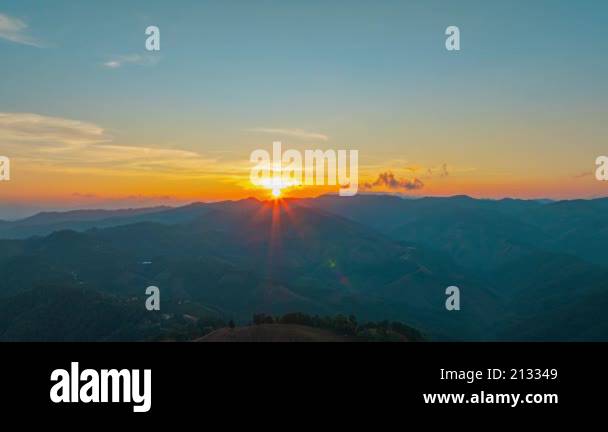 The sun sets behind a dense forest, casting a golden glow through ...