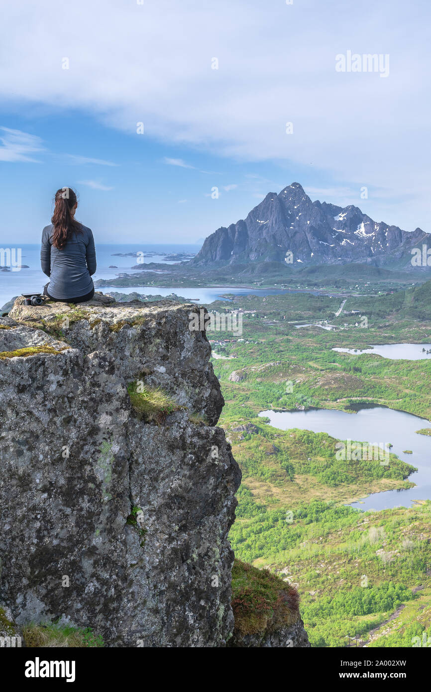 An attractive woman climbs to the top of a mountain to take in the amazing scenery above the towns of Kabelvag and Svolvaer in the Lofoten Islands. Stock Photo