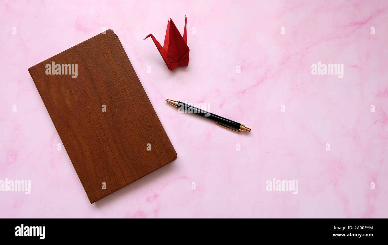A notebook with wooden cover, with a ball pen and a red paper crane beside it. Pink marble background. Stock Photo