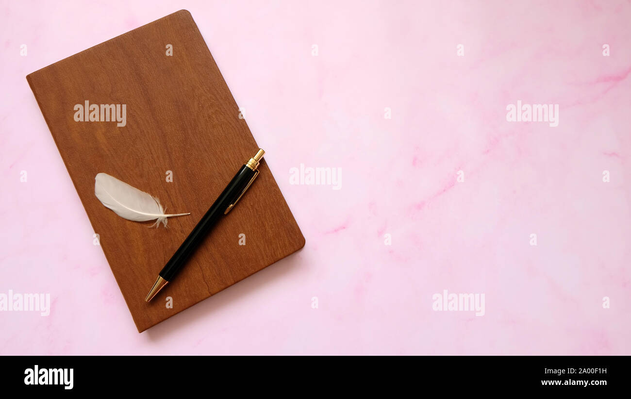 A notebook with wooden cover, with a ball pen and a white feather on top of it. Pink marble background. Stock Photo