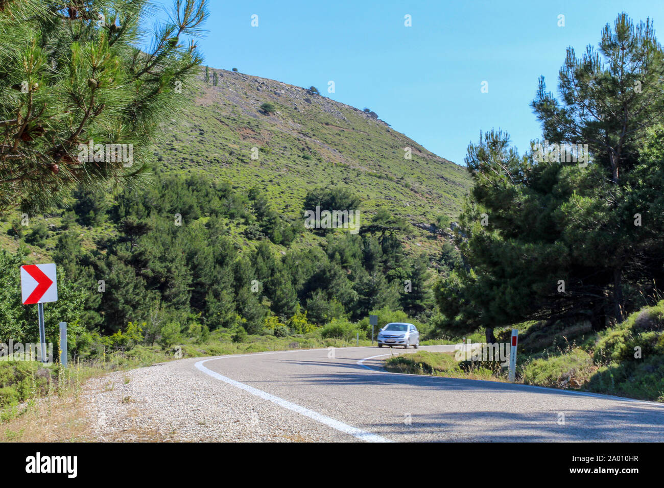 Car on asphalt island road, about to  cornering. Road lined with trees and mountains around Stock Photo