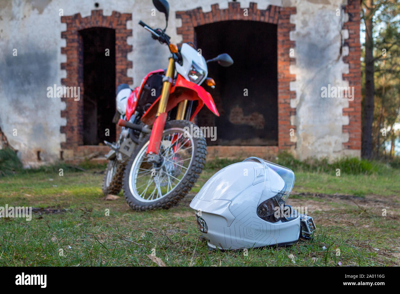 A white motorcycle helmet on the grass ground and a red motorcycle in the back, standing in front of the concrete mountain hut Stock Photo