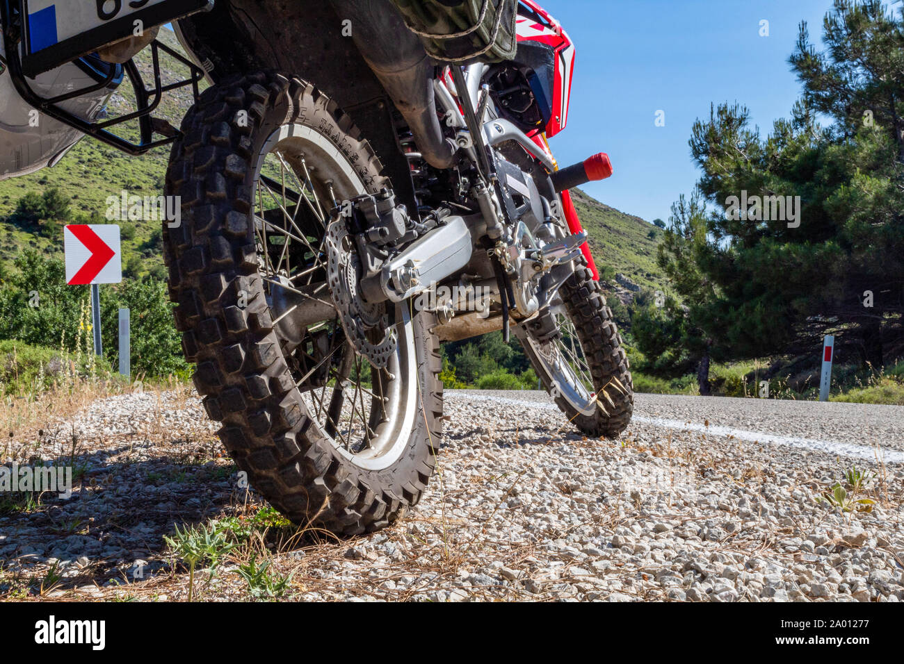 A red enduro motorcycle parked  on the gravel road on the side of the asphalt road. Only gear tire and mechanical parts are in the frame Stock Photo