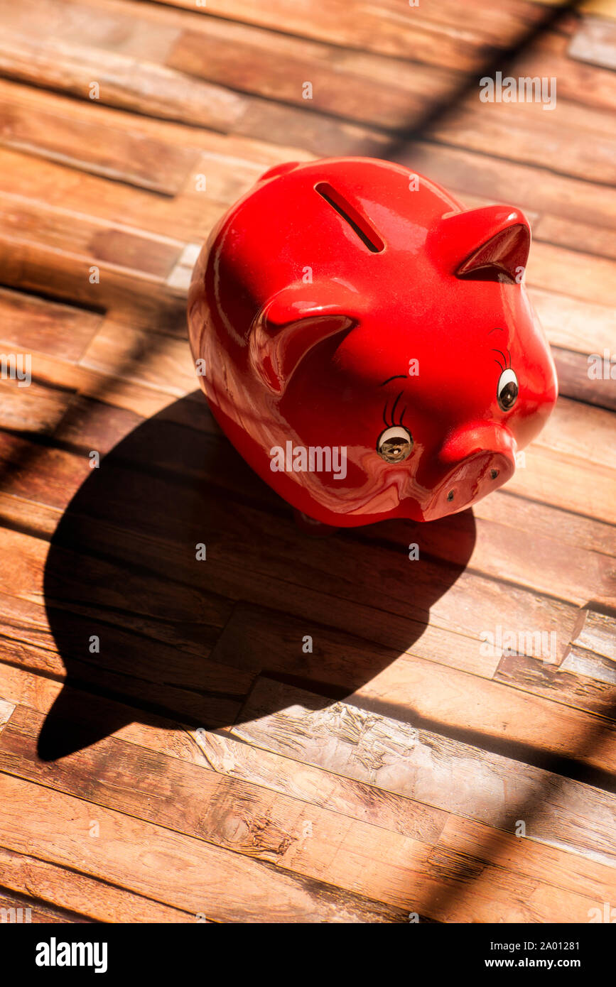 A red piggy bank stands on a wooden floor and casts a strong shadow Stock Photo
