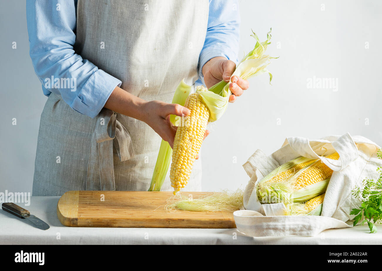 A female cook a cotton apron cleans ripe corn on the cob. Rustic natural style. The concept organic food. Kinfolk style. Stock Photo