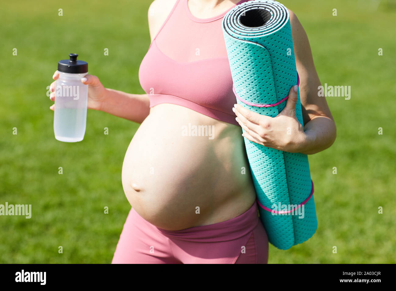 Close-up of pregnant woman in pink top holding exercise mat and bottle of water while standing outdoors after sports training Stock Photo