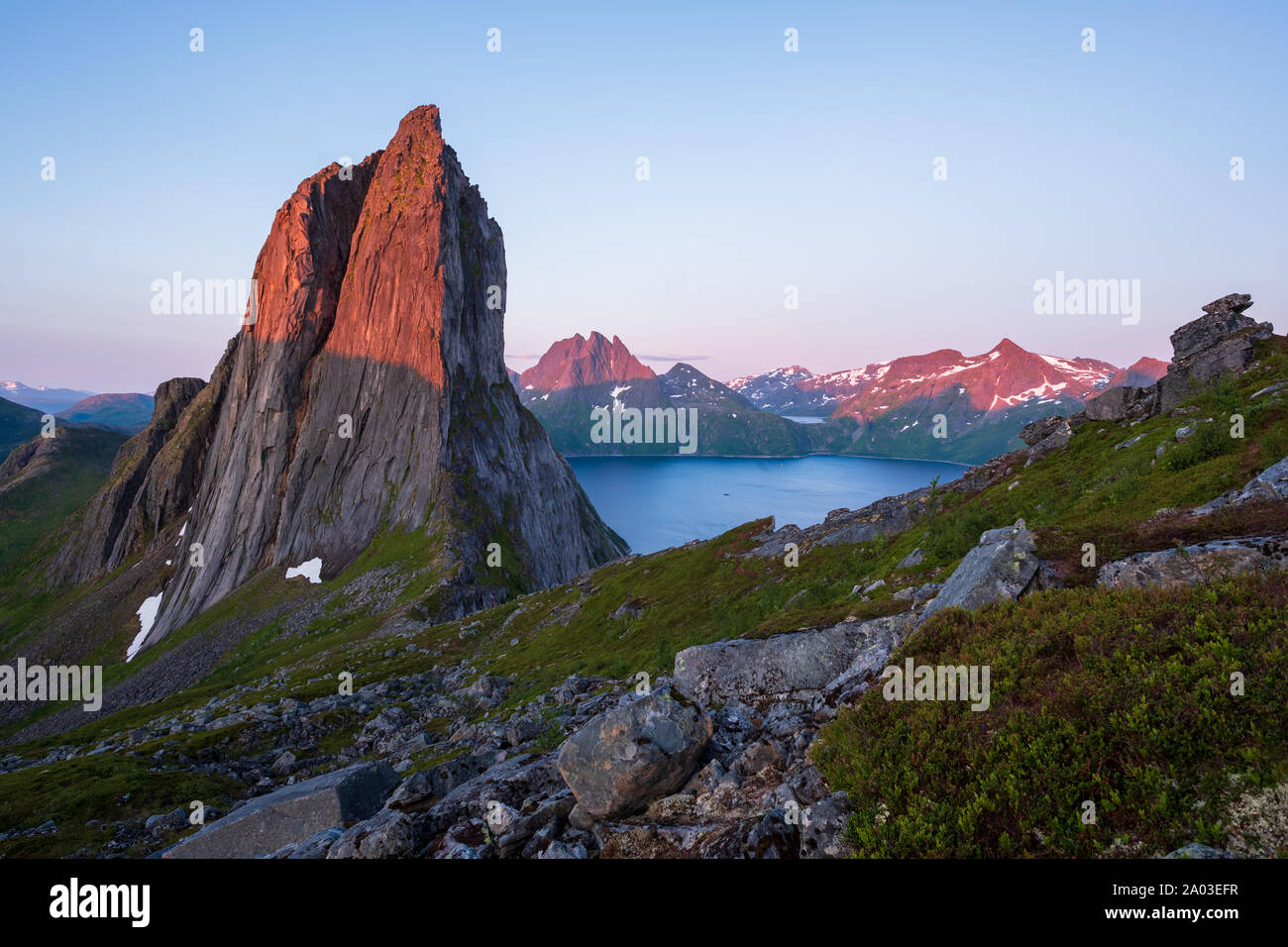 View from Mount Hesten on Iconic Mountain Segla in light of midnightsun in front of clear sky and mountain range in background, rocks and boulders in Stock Photo