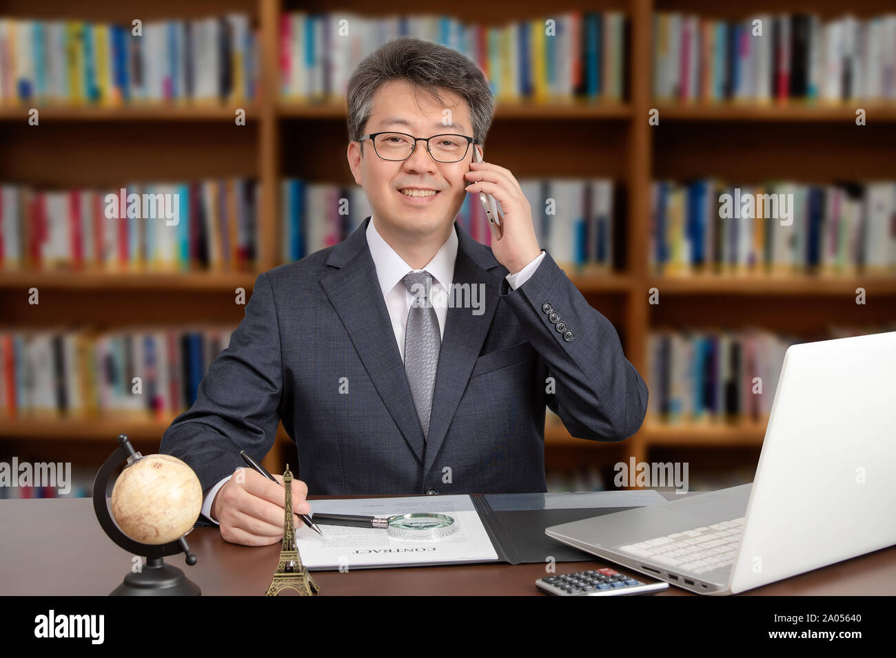 A portrait of an Asian middle-aged male businessman sitting at a desk, smiling and talking on the phone. Stock Photo