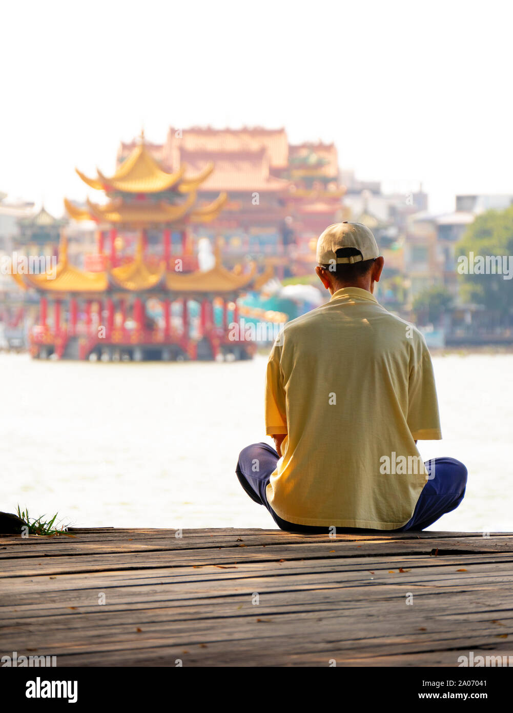 Man sitting in front of the Lotus Lake meditating. Chinese pagoda and temple in background. Lotus Pond Kaohsiung, Taiwan. Stock Photo