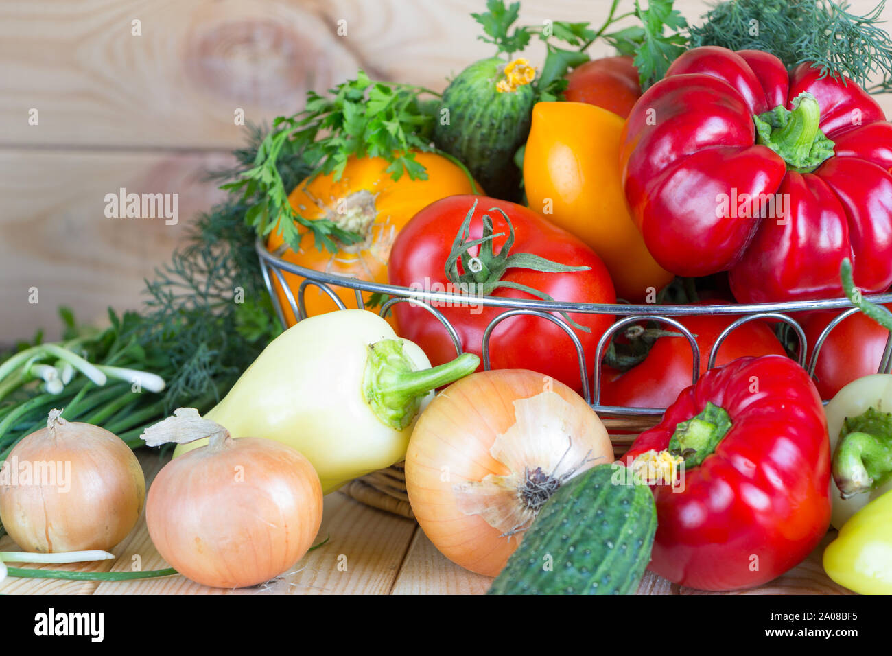 Close up vegetables harvest in wicker basket on wooden background. Red and yellow pepper, tomatoes, onion and green bow, parsley, dill and cucumbers. Stock Photo