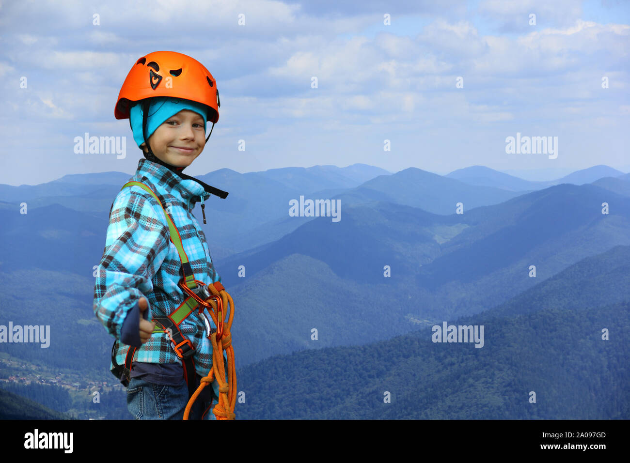 Portrait of a smiling six year old boy wearing helmet and safety harness in mountains, showing like symbol gesture, negative space Stock Photo