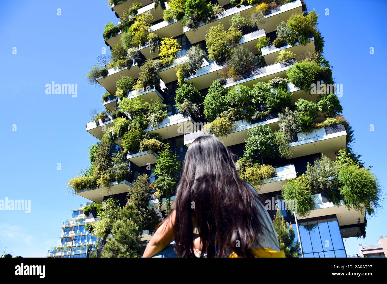 Beautiful thai tanned brunette is sitting in front of the Bosco Verticale (Vertical Forest) residential towers and looking up for the planted 900 tree Stock Photo