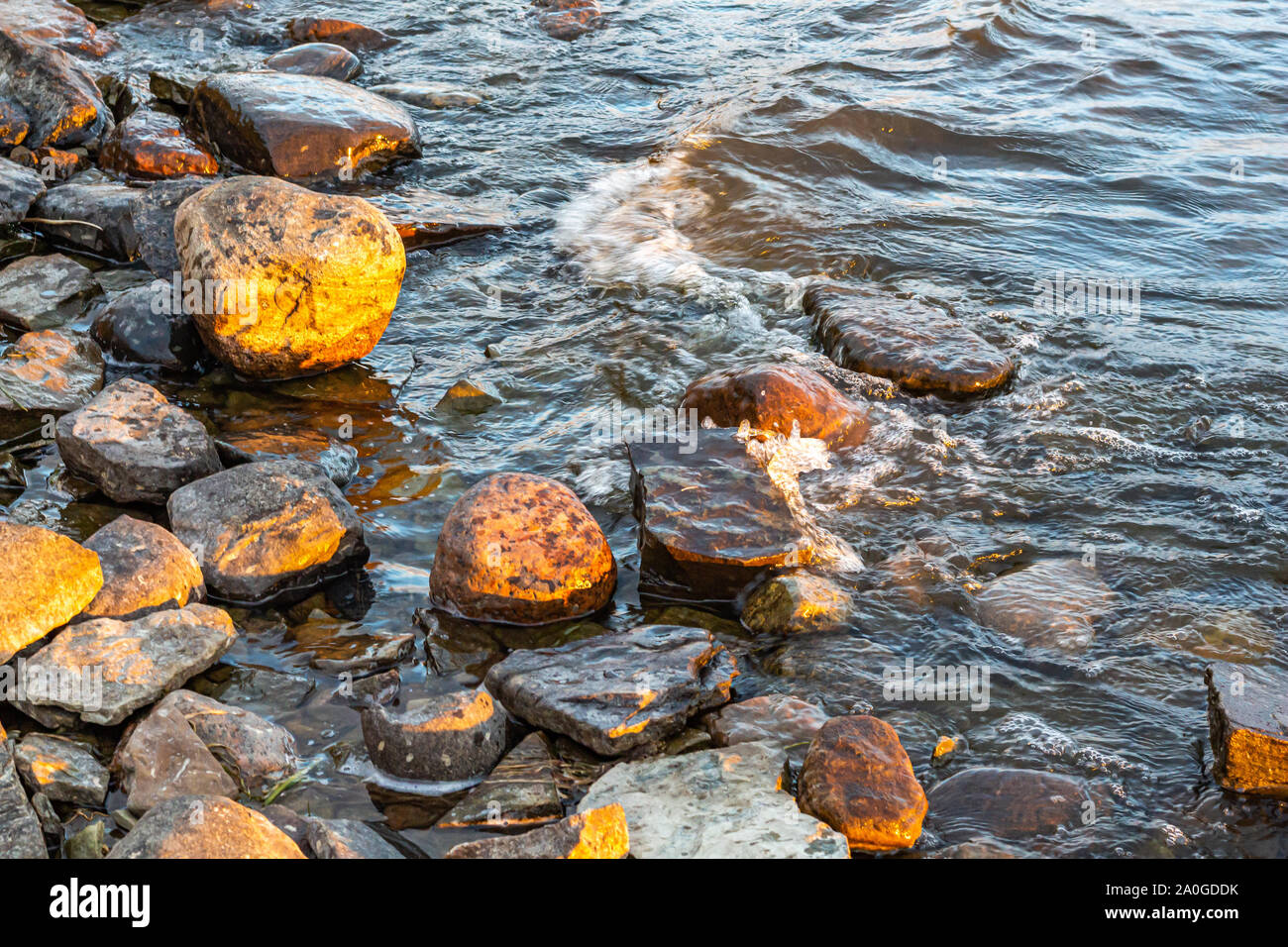 Small waves in a river roll into a rocky riverbank in the early morning light. Stock Photo