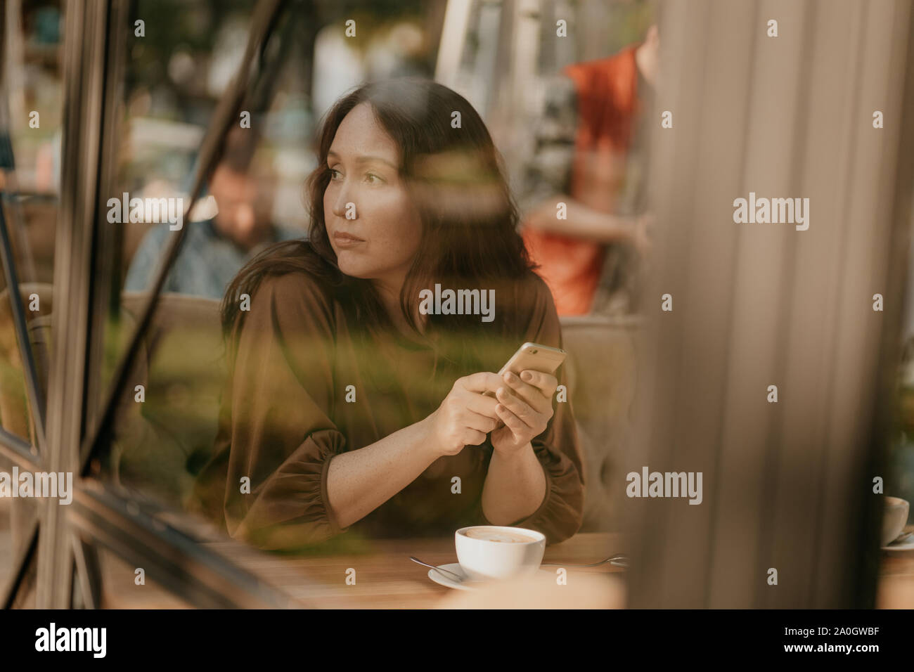Charming brunette woman with long curly hair sitting at the window in cafe with mobile phone in hands Stock Photo