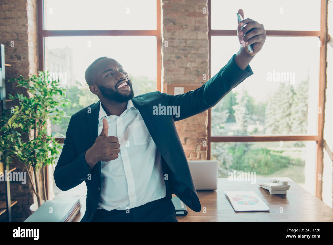 Elegant stylish classy cheerful positive business shark, mulatto man in jacket taking selfie on smartphone at workplace, workstation Stock Photo