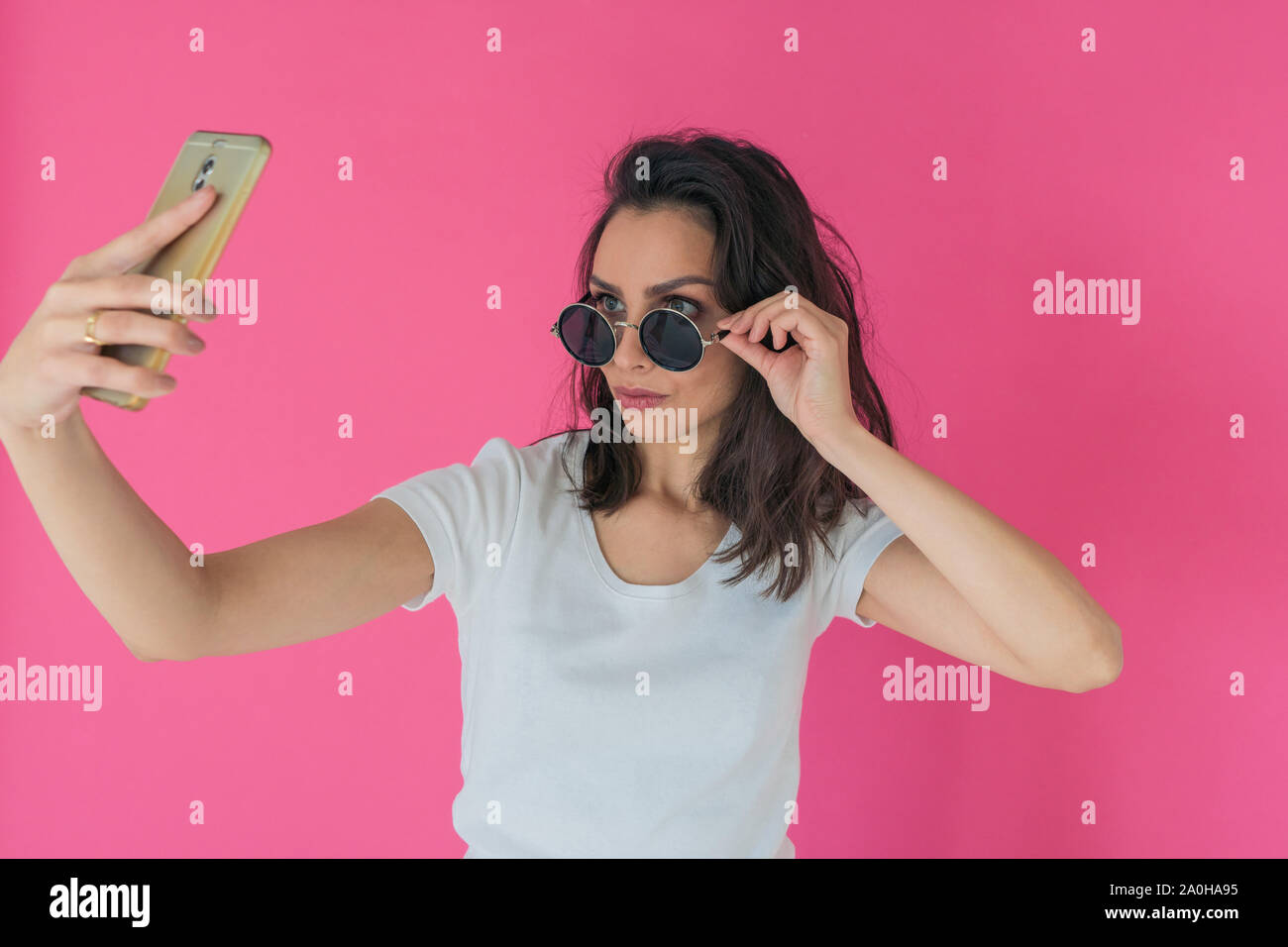 Beautiful girl in round sunglasses takes a selfie on the phone on a hot pink background Stock Photo