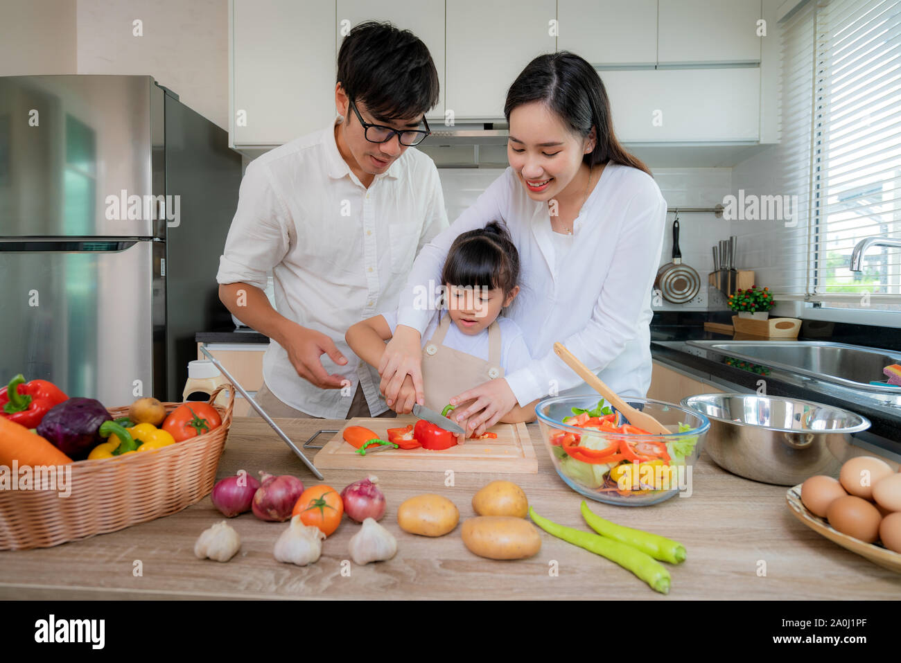 Asian mother teaching her daughter shredded vegetable salad with father standing on the side while the family was cooking in the kitchen at home. Fami Stock Photo