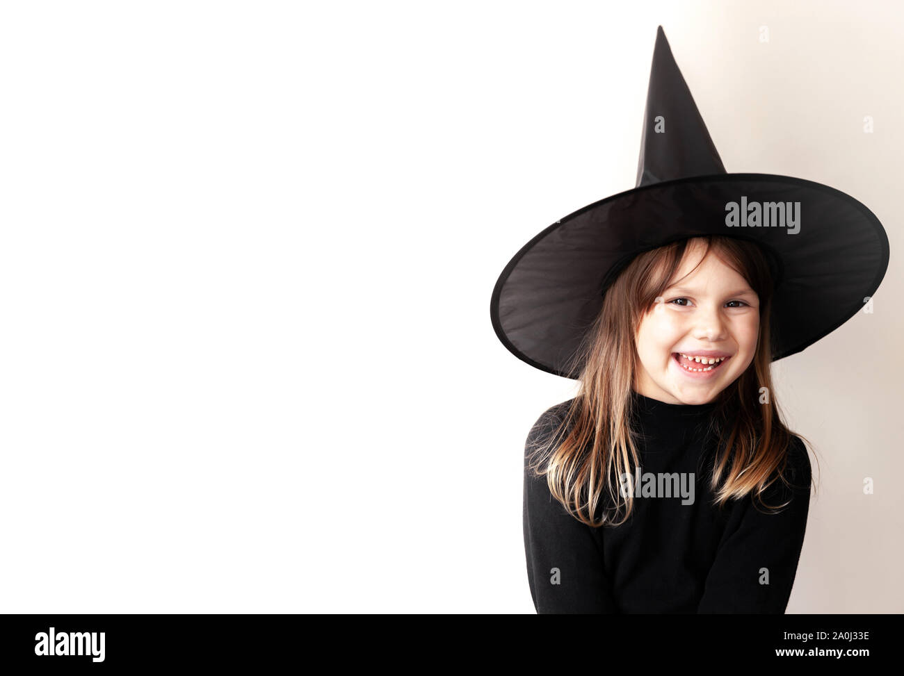 Little blond Caucasian girl in black witch costume smiles over white wall, studio portrait with copy space area on a left side Stock Photo