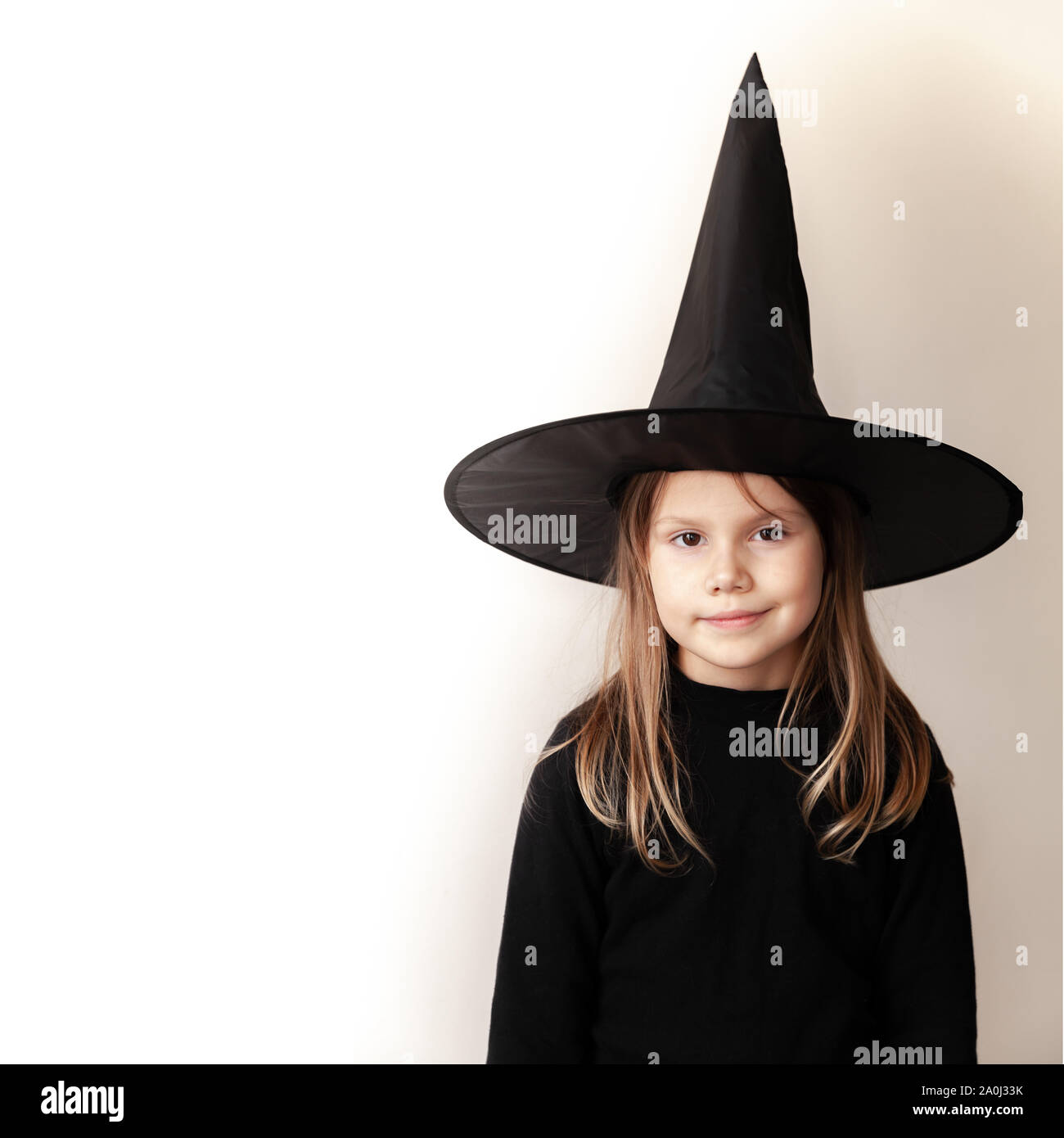 Little blond Caucasian girl in black witch costume smiles over white wall, close-up studio portrait with copy space area on a left side Stock Photo