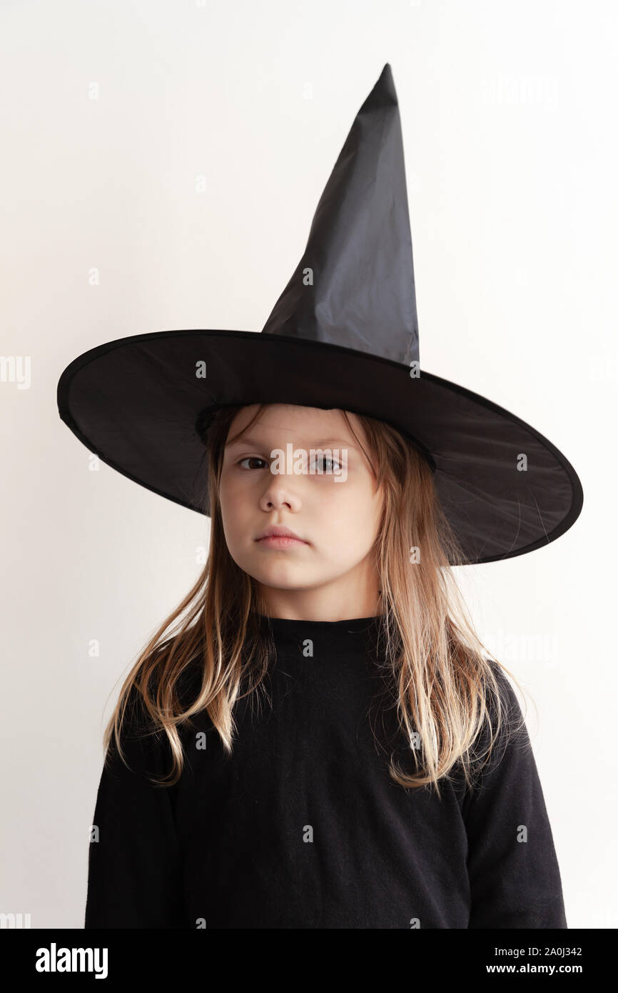 Little blond European girl in black witch costume with hat over white wall Stock Photo
