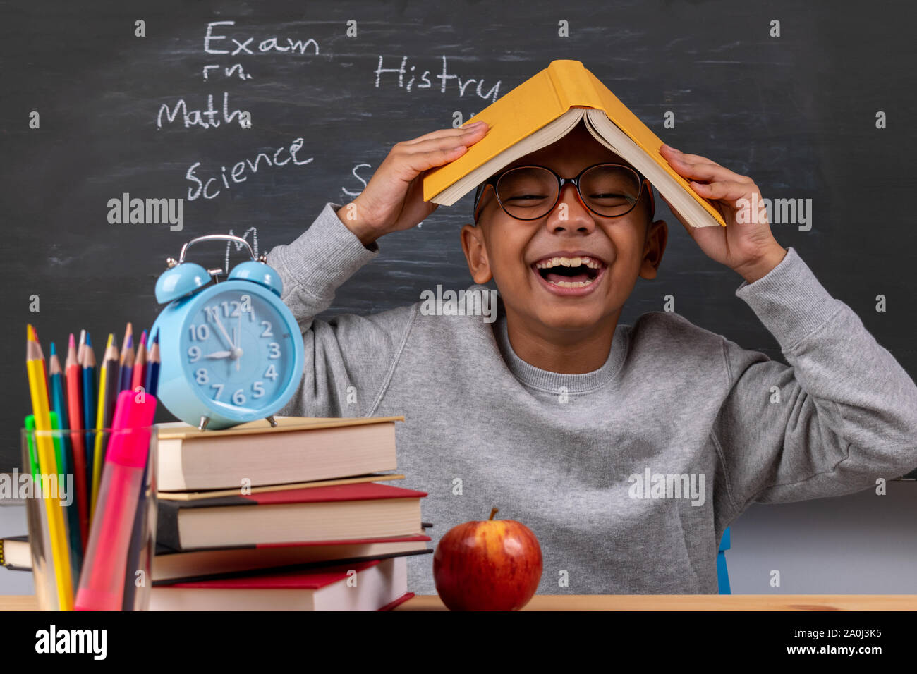 Cheerful thoughtful school boy, back to school concept. Stationery supplies on classroom desk with chalkboard background. Stock Photo