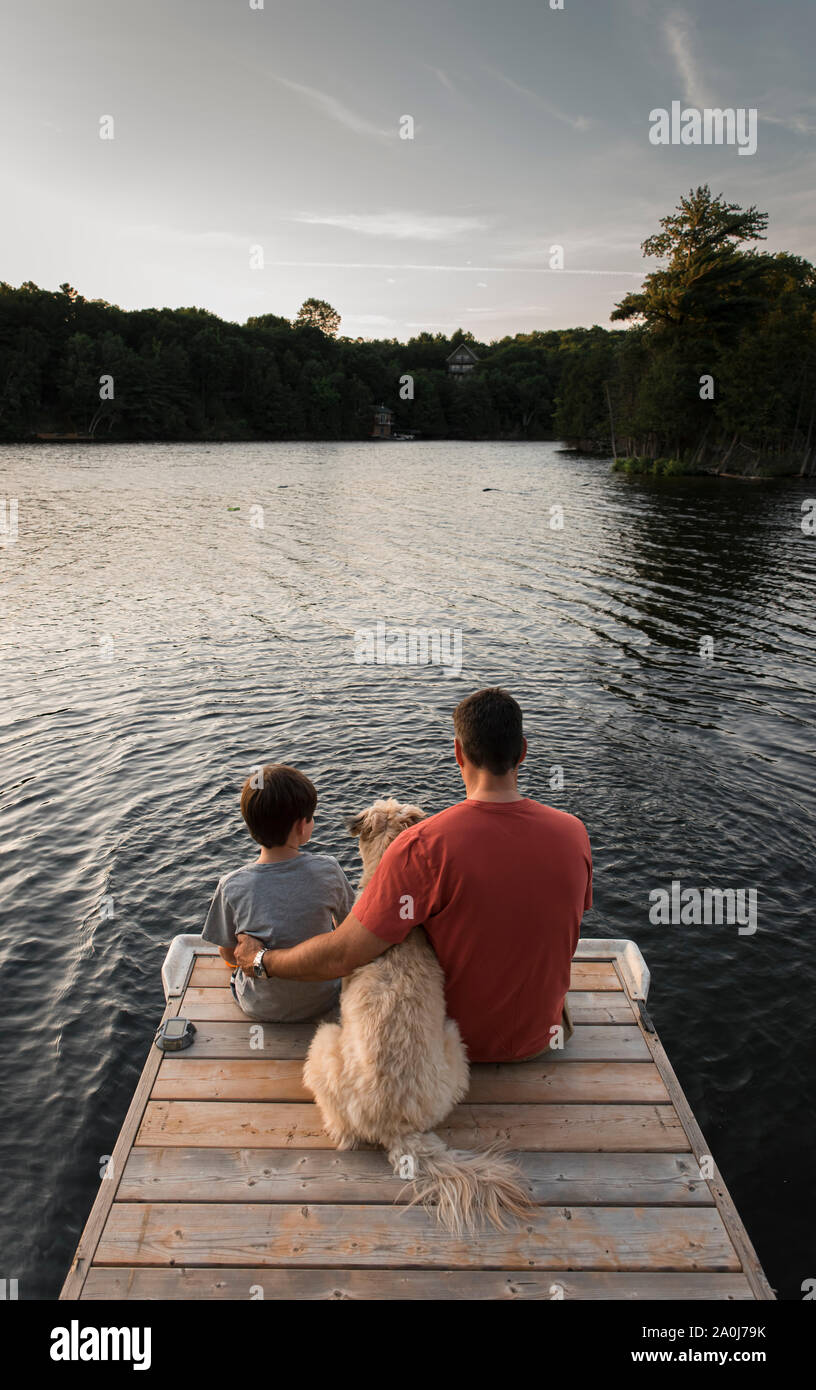Father and son sitting on end of a dock on a lake with their dog. Stock Photo
