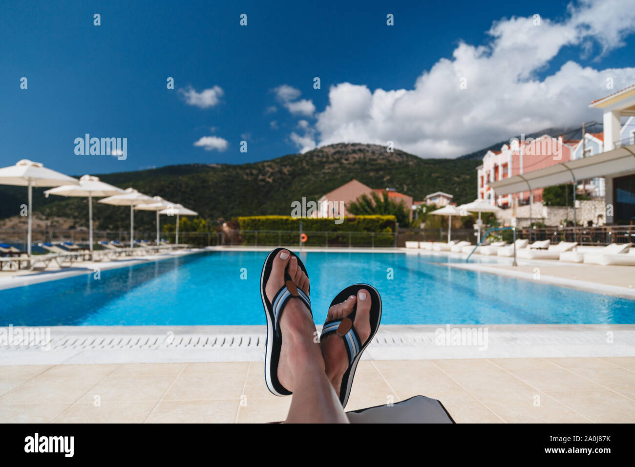 Close up view of young woman feet in blue flip-flops lying on a sun lounger with a swimming pool and blue sky at the background. Kefalonia island Stock Photo