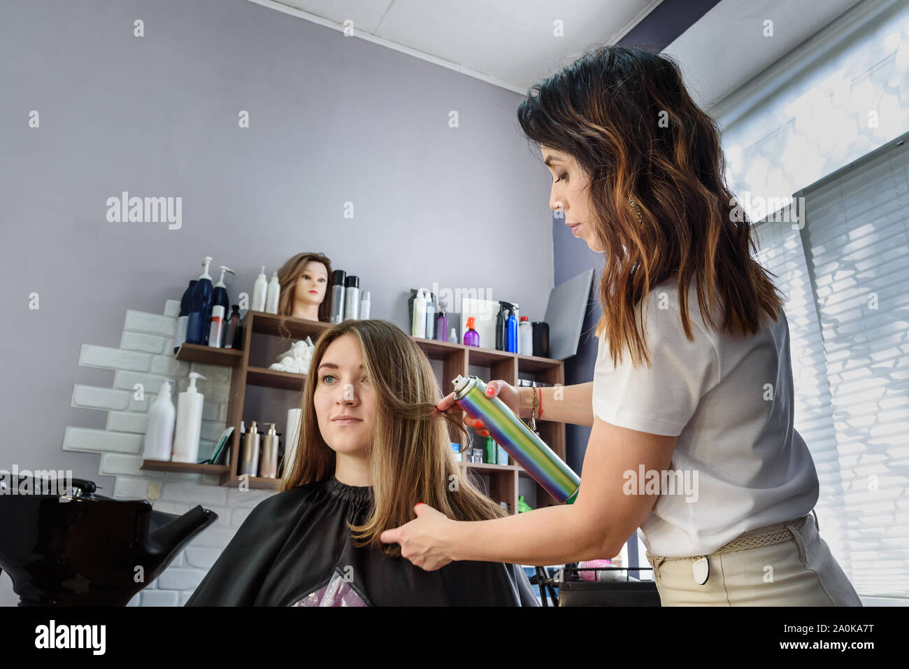 Professional hairdresser girl using hairspray on long brown hair. Process of hair styling in beauty salon Stock Photo