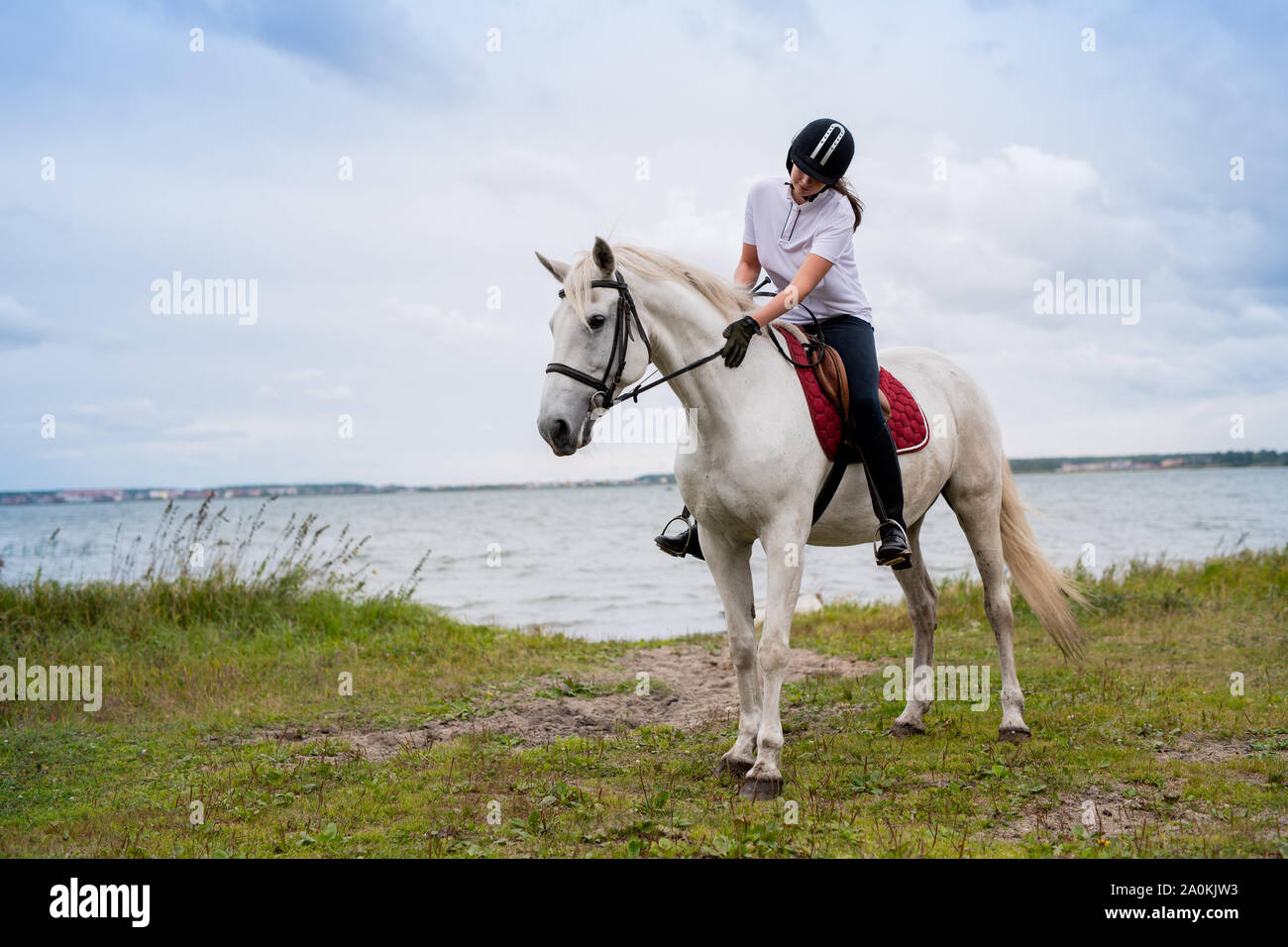 Active woman in equestrian outfit moving along river on back of white racehorse Stock Photo