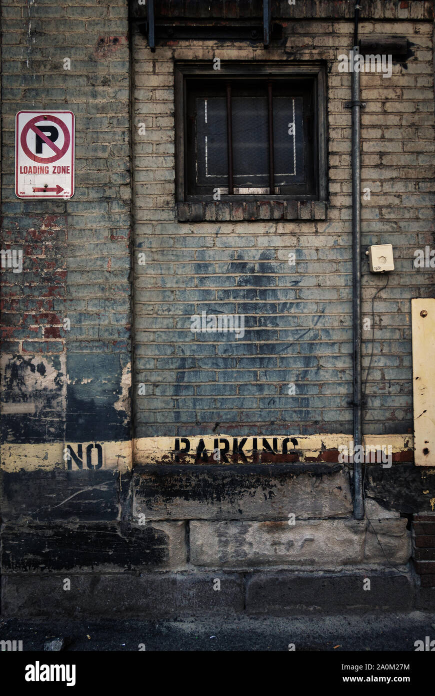 A weathered brick wall in an alleyway featuring a no parking sign a barred basement window and peeling paint with a gritty urban aesthetic Stock Photo