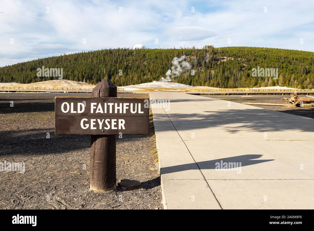 Old Faithful Geyser sign at Yellowstone National Park with steam emitting from the geyser in the background. Stock Photo