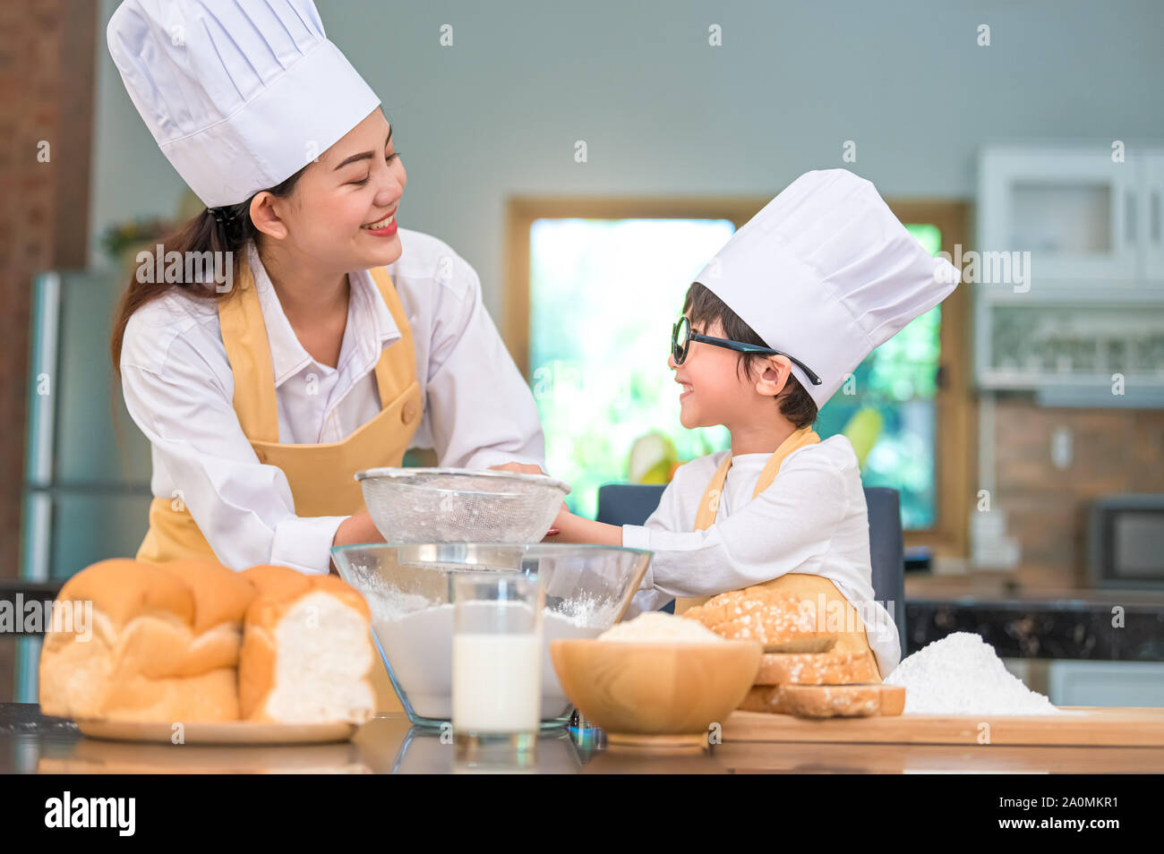 Cute little Asian boy and beautiful mother sifting dough flour with sifter sieve colander in home kitchen on table for prepare to baking bakery and ca Stock Photo